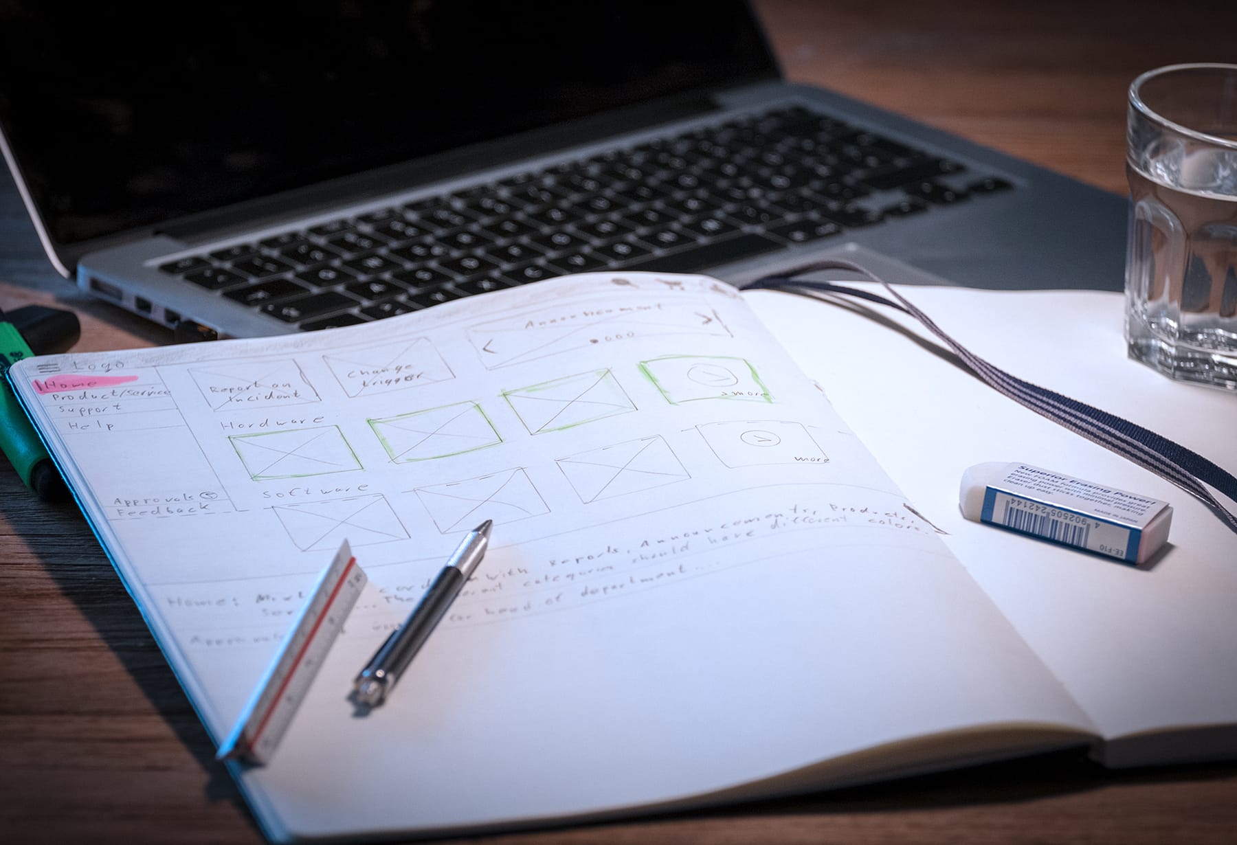 Shows a pad of paper with scribbles from a website, a ruler and pens on it. In the background, a blurry notebook and, to the right, a partially visible glass of water.